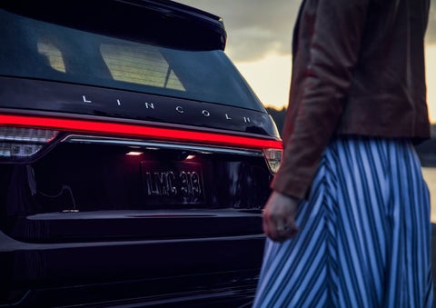 A person is shown near the rear of a 2024 Lincoln Aviator® SUV as the Lincoln Embrace illuminates the rear lights | Lincoln Demo 5 in Derwood MD