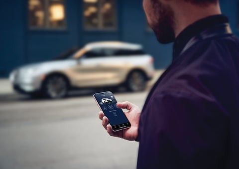 A person is shown interacting with a smartphone to connect to a Lincoln vehicle across the street. | Lincoln Demo 5 in Derwood MD